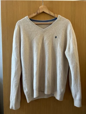 Beige V-ringad tröja med brodyr - Beige V-ringad tröja med diskret blå brodyr på bröstet. Tröjan har långa ärmar och ribbade muddar vid ärmslut och nederkant. Tillverkad i mjukt material som känns skönt mot huden. Enkel och stilren design som passar till det mesta.