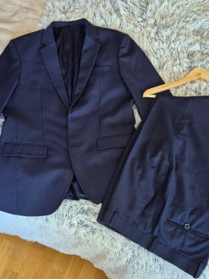 Mörkblå kostym med kavaj och byxor - Snygg mörkblå kostym med klassisk kavaj och matchande byxor. Kavajen har två knappar, smala slag och fickor med lock. Perfekt för dig som vill ha en stilren och tidlös look. Materialet känns mjukt och följsamt.