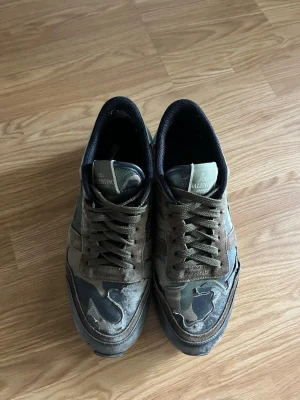 Valentino camo sneakers - Säljer ett par snygga sneakers från Valentino med camouflage-mönster i grönt, brunt och svart. Skorna har låga skaft, gröna snören och detaljer i mocka och läder. Perfekta för dig som gillar streetwear och vill sticka ut med exklusiva skor.