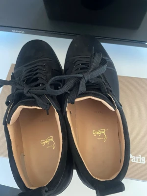 Svarta Louboutin sneakers i mocka - Snygga svarta sneakers från Christian Louboutin i mocka med klassisk röd sula och svarta skosnören. Låg modell med diskret logga på insidan och stilren design. Perfekta för dig som vill ha något exklusivt och trendigt.