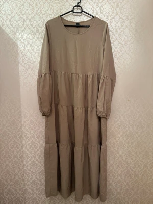 Beige långklänning med volang - Långklänning i beige med rund halsringning och långa puffärmar. Klänningen har flera volangdetaljer längs kjolen som ger en luftig och avslappnad look. Tillverkad i ett mjukt bomullsliknande material, perfekt för en trendig och bekväm stil.