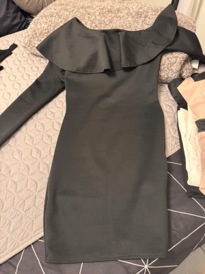 Svart oneshoulder klänning Glamorous - Snygg svart oneshoulder klänning från Glamorous med volangdetalj över axeln. Klänningen är kort och har en lång ärm på ena sidan, vilket ger en trendig och unik look. Perfekt för dig som vill sticka ut lite extra.