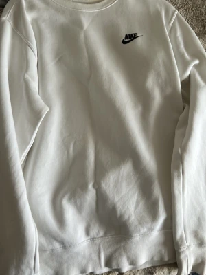 Vit sweatshirt från Nike - Säljer en stilren vit sweatshirt från Nike med klassisk svart logga på bröstet. Tröjan har rund halsringning och långa ärmar. Perfekt för en clean och sportig look.