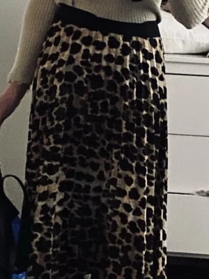 Leopardmönstrad långkjol - Säljer en snygg långkjol med leopardmönster och bred svart midjeresår. Kjolen har ett plisserat fall och är perfekt för dig som vill sticka ut lite extra. Passar till både stickad tröja och t-shirt.