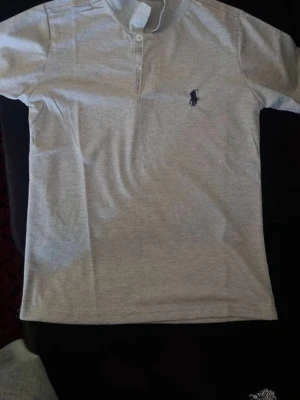 Grå t-shirt från Polo Ralph Lauren - Snygg grå t-shirt från Polo Ralph Lauren med klassisk pikékrage och knappslå. T-shirten har korta ärmar och den ikoniska loggan broderad på bröstet. Perfekt för en stilren och avslappnad look.