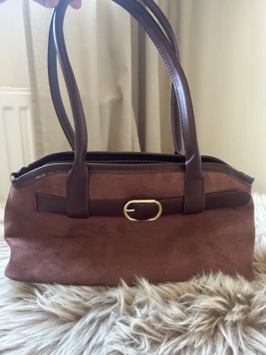 Brun mocka handväska med guldspänne - Snygg handväska i brun mocka med matchande mörkbruna handtag och dekorativt guldspänne framtill. Perfekt accessoar för dig som gillar klassisk och stilren design. 