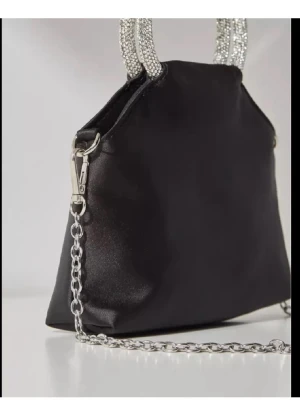 Svart kuvertväska med Primark - Snygg svart kuvertväska från Primark i satinliknande material. Väskan har glittrande handtag täckta av strass och en avtagbar silverfärgad kedja som axelrem. Perfekt för att lyfta din outfit med lite extra bling.