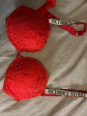 Röd spets-bh från Victoria's Secret - Snygg röd bh från Victoria's Secret med vadderade kupor och spetsdetaljer. Axelbanden är dekorerade med glittrande stenar och texten 'Victoria's Secret'. Perfekt för dig som vill ha något extra fint under kläderna. Vet inte exakt storlek eftersom att lappen ibte sitter kvar men tror det är 75 B/C