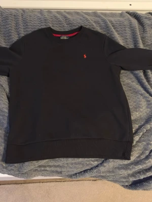 Svart sweatshirt från Polo Ralph Lauren - XL men de är S , Klassisk svart sweatshirt från Polo Ralph Lauren med rund halsringning och den ikoniska röda logotypen broderad på bröstet. Tröjan har ribbade muddar vid ärmslut och nederkant. Perfekt för en clean och stilren look.
