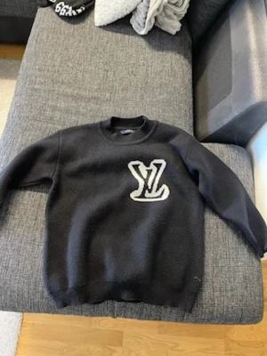 Svart Louis Vuitton tröja - Säljer en svart tröja från Louis Vuitton med stor vit LV-logga på bröstet. Tröjan har rund halsringning och ribbade muddar vid ärmslut och nederkant. Perfekt för dig som gillar exklusiva streetwear-plagg.