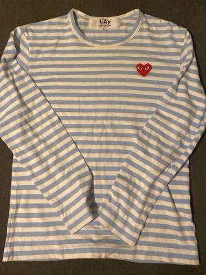 Randig långärmad topp från Comme des Garçons - Säljer en långärmad topp från Comme des Garçons Play med ljusblå och vita ränder. Toppen har ett ikoniskt rött hjärta broderat på bröstet och är gjord i mjuk bomull. Perfekt för dig som gillar stilrena och trendiga plagg.