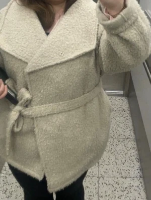 Beige teddyjacka med knytskärp - Säljer en supermjuk och fluffig beige teddyjacka med bred krage och knytskärp i midjan. Jackan har en oversized passform och stora slag, perfekt för att hålla värmen under kyliga dagar. Materialet är klassiskt teddytyg som ger en mysig look.