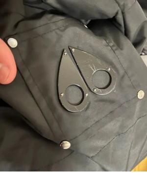 Svart dunjacka från Moose Knuckles - Säljer en svart dunjacka från Moose Knuckles med ikoniska metall-loggan på ärmen och broderad insida med hockeymotiv. Jackan har silverfärgade nitar och är fylld med dun för extra värme. Perfekt för kalla dagar och riktigt snygg streetstil.