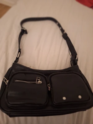 Svart axelväska med fickor - Cool svart axelväska i slätt material med två stora framfickor, en med dragkedja och en med tryckknappar. Justerbar axelrem med spänne. Perfekt för att bära det viktigaste och ger en streetstyle-vibe.