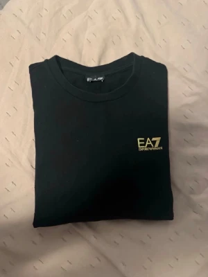 EA7 Emporio Armani sweatshirt  - Snygg svart t-shirt från EA7 Emporio Armani med gul logga på bröstet. Klassisk rund halsringning och stilren design som passar perfekt till jeans eller shorts. Tillverkad i mjuk bomull för skön känsla hela dagen.