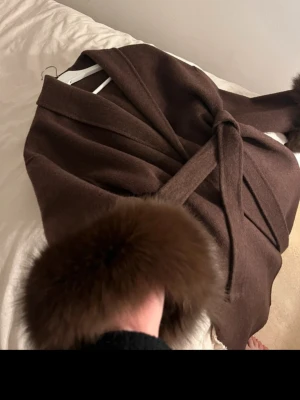 Brun ullkappa med pälskant - Snygg brun kappa i ull med bred krage och bälte i midjan. Ärmarna har lyxiga pälskanter som ger en exklusiv känsla. Perfekt för dig som vill ha en elegant och trendig look under kyliga dagar.