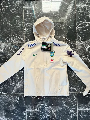Vit Brasilien Nike vindjacka - Snygg vit vindjacka från Nike med Brasilien CBF-logga och flera sponsortryck. Jackan har huva med snörning, dragkedja framtill och är tillverkad i GORE-TEX-material. Perfekt för dig som vill ha sportig stil och gillar fotboll.