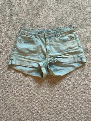 Ljusblå denimshorts med uppvik - Säljer ett par ljusblå denimshorts med klassisk femficksmodell och uppvikta benslut. Shortsen har bälteshällor, knapp och dragkedja framtill samt bakficka. Perfekta för sommaren och riktigt sköna att bära.