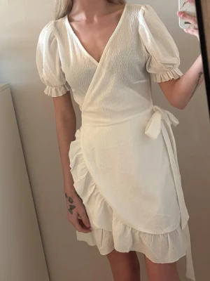 Vit omlott klänning - Fin omlott klänning med ett snöre runt midjan. Volanger på kanterna. Aldrig använd endast testad. Inga defekter. Perfekt för student eller andra vardagliga tillfällen! 