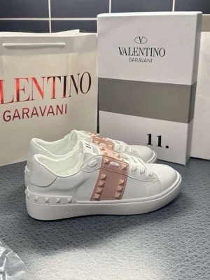 Valentino Garavani Rockstud sneakers - Snygga vita sneakers från Valentino Garavani med ljusrosa band över vristen dekorerat med ikoniska nitar. Skorna har en klassisk låg siluett och är tillverkade i slätt läder med vita snören och detaljerade nitar runt hälen. Perfekta för dig som gillar exklusiv och edgy stil.