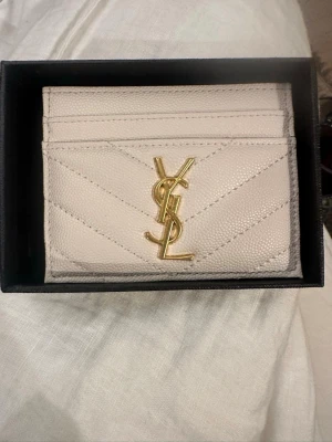 Beige korthållare från Saint Laurent - Snygg beige korthållare från Saint Laurent i läder med guldfärgad YSL-logga framtill. Plånboken har flera kortfack och ett elegant, quiltat mönster. Perfekt accessoar för dig som gillar lyxiga detaljer och stilren design. Ej äkta