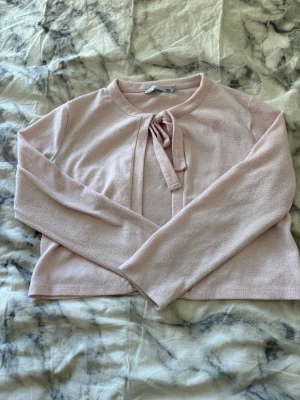 Ljusrosa kofta med knytband - Söt ljusrosa kofta med långa ärmar och knytband framtill vid halsen. Koftan har en mjuk, lite fluffig textur och en enkel, rak passform. Perfekt att slänga över en topp för en chill och stilren look.