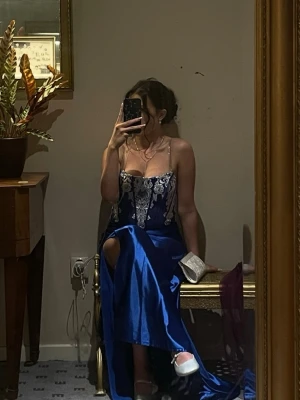 Blå långklänning med spetsdetaljer - Säljer en elegant blå långklänning med vit spets upptill. Klänningen har en slits på sidan och figurnära passform. Perfekt för bal eller festliga tillfällen. Materialet är glansigt och ger en lyxig känsla.