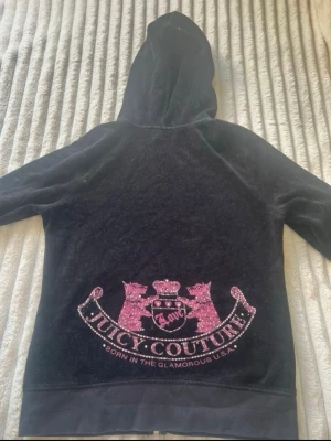 Juicy Couture svart velour hoodie - Juicy Couture hoodie i svart velour med huva. Baksidan har ett stort rosa glittrigt emblem med två hundar, krona och texten 'Juicy Couture' samt 'Born in the Glamorous U.S.A.