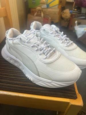 Vita sneakers från Puma - Snygga vita sneakers från Puma med modern design och detaljer i mesh och syntet. Skorna har vita snören med grå inslag och en robust sula med mönster för bra grepp. Perfekta för dig som gillar stilrena och sportiga skor.