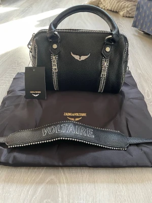 Svart handväska Zadig & Voltaire - Snygg svart handväska i strukturerat läder från Zadig & Voltaire. Väskan har silverfärgade detaljer, dubbla dragkedjor framtill och ett emblem med vingar. Axelremmen är dekorerad med texten VOLTAIRE och nitar längs kanten. Perfekt accessoar för en trendig look.