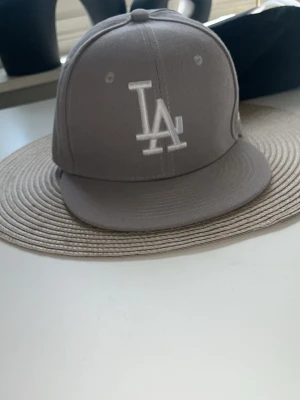 Grå LA New Era keps - Snygg grå snapback-keps från New Era med LA-logga broderad i vitt framtill. Klassisk rak skärm och ventilerande hål upptill. Kepsen är tillverkad i bomullsmix och har en stilren look som passar till streetwear och casual outfits.