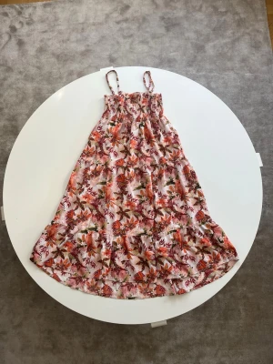 Blommig sommarklänning med smala axelband - Söt blommig klänning med smala axelband och rynkad överdel. Klänningen har ett färgglatt mönster med olika sommar färger🌸. Klänningen är i storlek 134 och är perfekt för varma dagar och har en luftig, avslappnad passform.💕