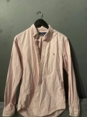 Randig skjorta från Polo Ralph Lauren - Säljer en slim fit skjorta från Polo Ralph Lauren i vitt och rött med tunna ränder. Den är knappast använd så den är i ny skick, skulle bara kanske behöva en strykning. Kontakta gärna vid intresse:)