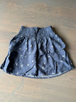 Mörkblå blommig omlottkjol - Supersöt mörkblå kortkjol med små vita och rosa blommor. Kjolen har bred smock i midjan och omlottdesign som ger en avslappnad och trendig look. Perfekt för varma dagar och enkel att matcha med en enkel topp.