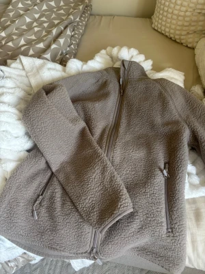 Beige fleece jacka med dragkedja - Mysig beige teddyjacka med hög krage och hel dragkedja framtill. Jackan har två sidofickor med dragkedja och är gjord i fluffigt teddy-material som håller dig varm. Perfekt för kyliga dagar och enkel att matcha med olika outfits.