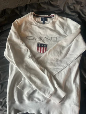 Vit sweatshirt från GANT - Säljer en vit sweatshirt från GANT med broderad logga och ett sköldmärke med stjärnor och ränder på bröstet. Tröjan har rund halsringning, långa ärmar och ribbade muddar. Perfekt för en clean och klassisk stil. Aldrig använd
