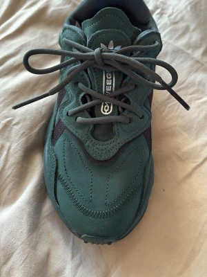 Adidas grön sneaker Ozweego - Snygga gröna Adidas Ozweego sneakers med svarta detaljer och klassiska tre ränder på sidan. Skorna har en chunky sula och är gjorda i mocka och mesh för en cool och sportig vibe. Perfekta för dig som vill sticka ut med stilrena och trendiga sneakers.