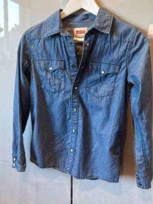 Blå jeansskjorta från Levi's - Snygg blå jeansskjorta från Levi's med klassisk krage, tryckknappar och två bröstfickor med lock. Skjortan har långa ärmar och är gjord i mjuk bomull. Perfekt för en avslappnad och cool stil.