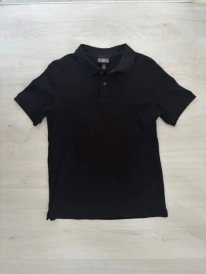 Svart pikétröja från H&M - Klassisk svart pikétröja från H&M med krage och två knappar framtill. Tröjan har korta ärmar och är tillverkad i mjuk bomull som känns skön mot huden. Perfekt till jeans eller shorts för en clean och enkel look.