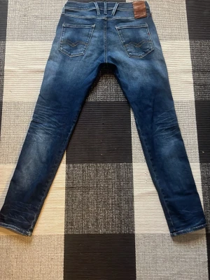 Replay anbass hyperflex - Snygga blå slim fit jeans från Replay med klassisk femficksdesign och coola slitningar. Jeansen har kontrastsömmar på bakfickorna och en brun läderpatch med Replay-logga i midjan. Perfekta för en trendig och avslappnad stil.