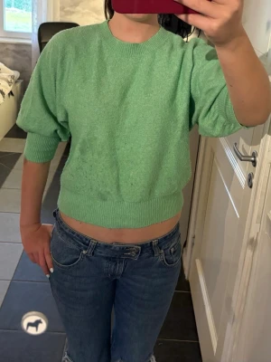 Grön kortärmad stickad tröja - Snygg ljusgrön stickad tröja med korta, puffiga ärmar och bred ribbad mudd i midjan och vid ärmsluten. Tröjan har en avslappnad passform och rund halsringning. Perfekt att matcha med jeans för en chill look.