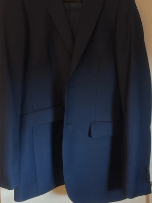 Marinblå kostym med kavaj och byxor - Snygg marinblå kostym med klassisk kavaj och matchande byxor. Kavajen har två framfickor med lock och knäppning framtill. Perfekt för dig som vill ha en stilren och tidlös look. Modellen är slim fit, kavajen strl 44 long och byxorna strl 20-32. Passar dig som är runt 180cm och vill ha en smal passform.