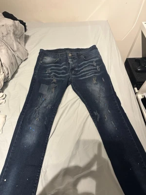 Mörkblå dsq2 jeans  - Säljer ett par mörkblå dsq2 jeans med slitningar, patchdetaljer och färgstänk. Jeansen har klassisk femficksmodell och knapgylf. Perfekta för dig som gillar en trendig och avslappnad streetstil.