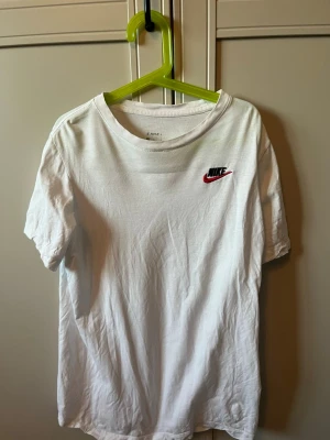 Vit Nike t-shirt med logga - Klassisk vit t-shirt från Nike med den ikoniska loggan i svart och rött på bröstet. T-shirten har rund halsringning och korta ärmar. Tillverkad i mjuk bomull som känns skön mot huden. Perfekt till en avslappnad och sportig stil.