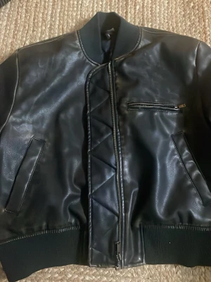 Svart skinnjacka från Divided XS - Snygg svart skinnjacka från Divided i storlek XS. Jackan har ribbade muddar, dragkedja framtill, två snedställda fickor och en bröstficka med dragkedja. Cool quiltad detalj längs dragkedjan och klassisk bomberjacke-look.