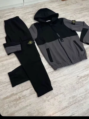 Stone Island loungewear set svart/grå - Tvådelat loungewear set från Stone Island i svart och grått. Setet består av en zip-hoodie med huva och matchande byxor. Båda delarna har Stone Island-logga på ärm och ben, samt praktiska fickor. Perfekt för chill dagar hemma eller när du vill vara bekväm.