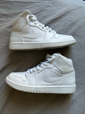 Nike Air Jordan 1 Mid vita sneakers - Snygga vita Nike Air Jordan 1 Mid sneakers med klassisk siluett och rund tå. Skorna har snörning och är tillverkade i skinn med diskreta detaljer och ikoniska loggor på sidan och skaftet. Perfekta för dig som gillar stilrena och tidlösa sneakers.