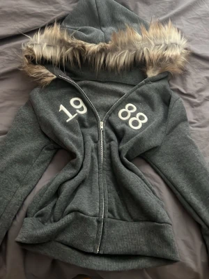 Grå hoodie med fuskpäls och dragkedja - Snygg 2000tals inspirerad grå hoodie med dragkedja och stor huva kantad med fuskpäls. Tryck med siffrorna 1988 på bröstet. Tillverkad i mjukt sweatshirtmaterial, perfekt för chill dagar. Ribbstickade muddar vid ärmslut och nederkant. 