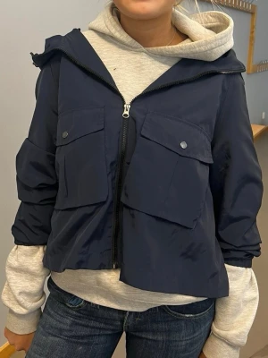 Mörkblå vindjacka med fickor - Snygg mörkblå vindjacka med stora framfickor och dragkedja framtill. Jackan har en lös passform och huva, perfekt för lager på lager. Passar till dig som gillar en avslappnad och sportig stil.