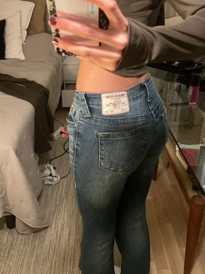 Blå bootcut jeans från True Religion - Säljer ett par blå bootcut jeans från True Religion med klassiska kontrastsömmar och stor bakficka med logga. Jeansen har en snygg tvättad look och är tillverkade i denim med lätt stretch. Perfekta för dig som gillar en avslappnad men trendig stil. orginal pris är 1299